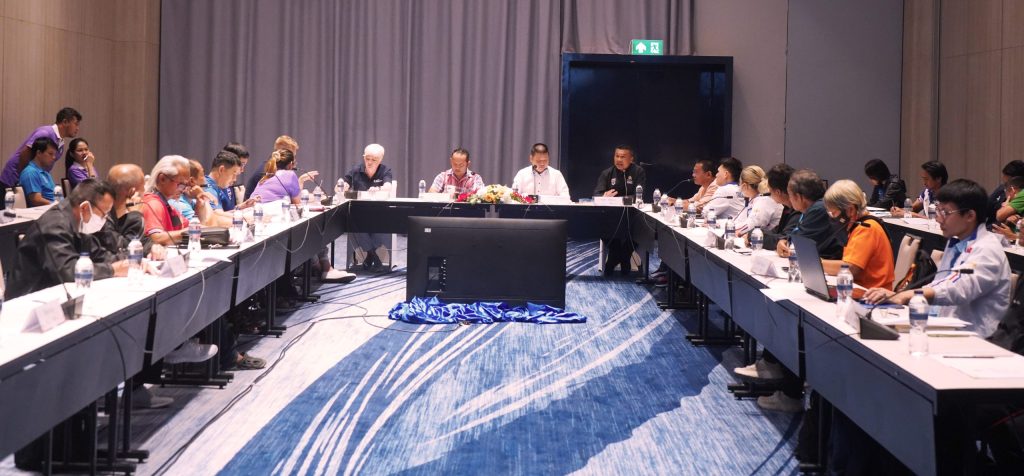 A meeting is pictured. A large number of people are sat around a table in a u-shape.