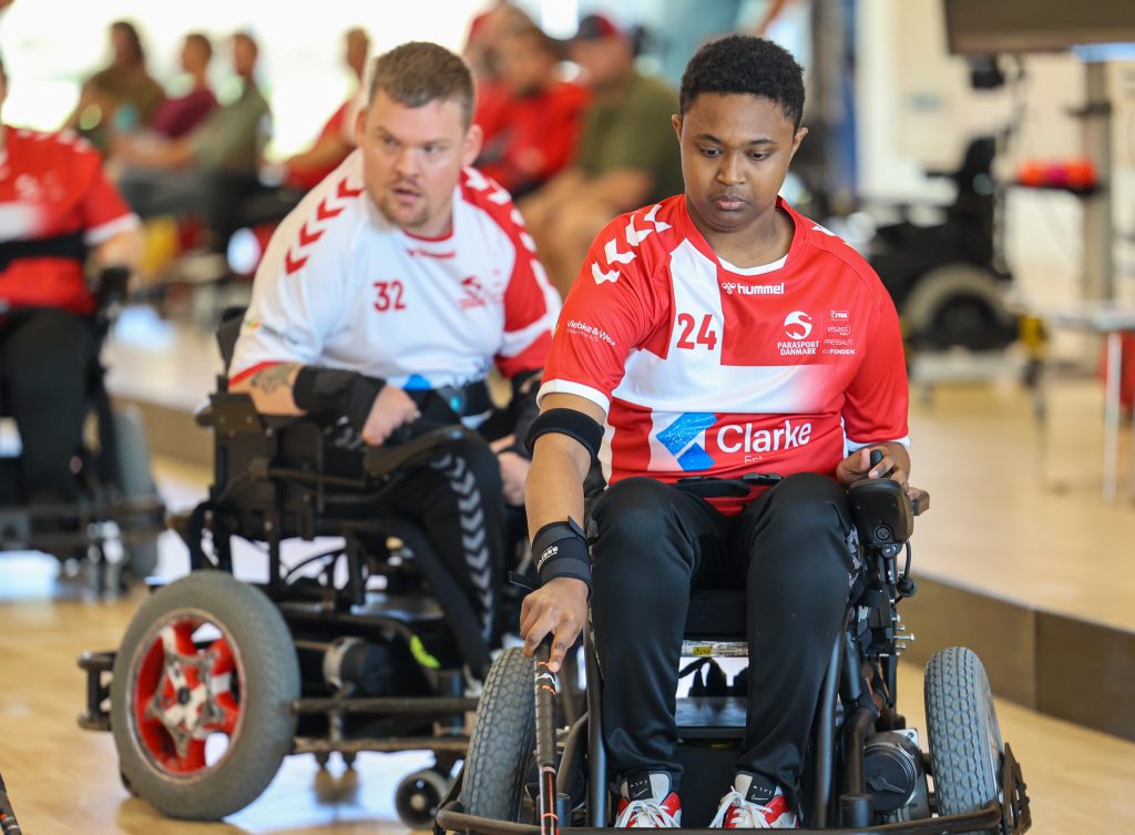 A powerchair hockey player from Denmark moves across the court closely followed by a player from an opposing team